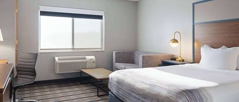 A hotel room with a large bed, a desk, a chair, a window, and a lamp.