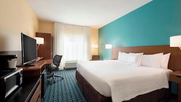 Una habitación de hotel con cama, escritorio con televisor, silla, lámpara y ventana con cortinas.