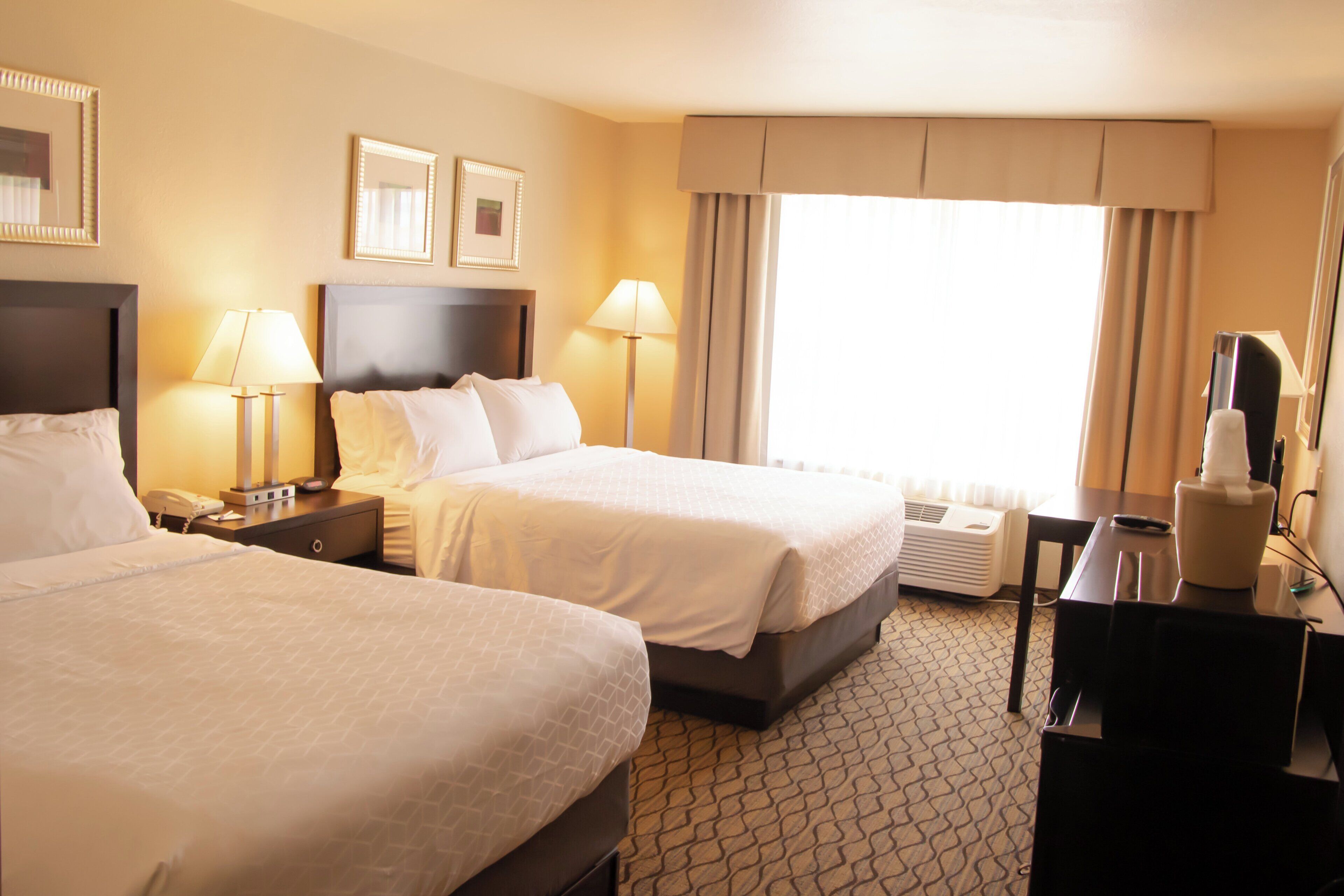 A hotel room with two beds, bedside lamps, a desk with a telephone, a TV, and a window with curtains.