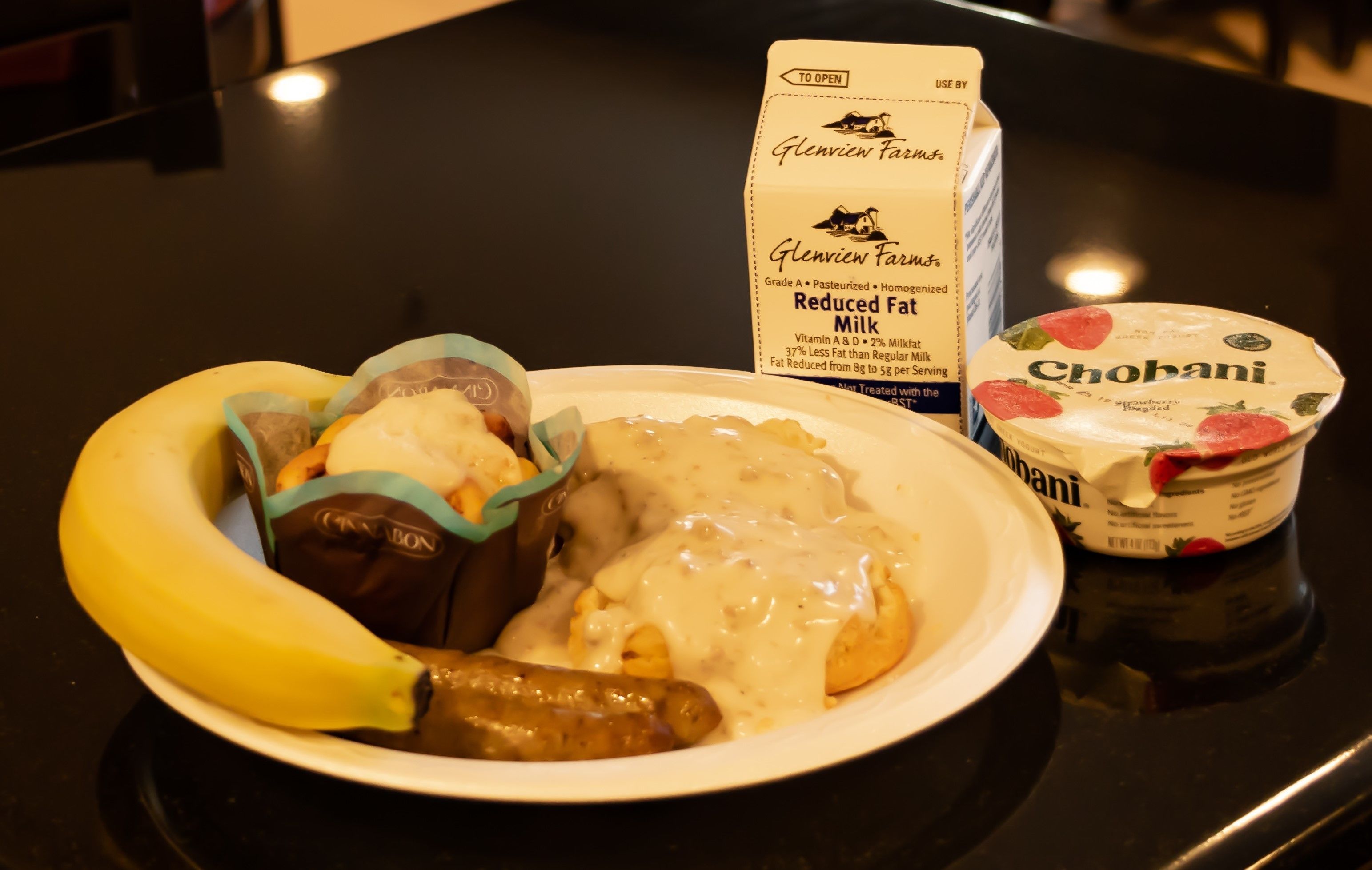 A plate with food items including a banana, sausage, and a dessert, alongside a carton of reduced fat milk and a tub of Chobani yogurt.