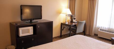 A hotel room with a bed, a television, a desk with a lamp, a chair, a microwave, and a waste bin.