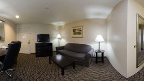 Habitación de hotel con sofá, una mesita, televisión en mueble bajo, escritorio con silla y dos lámparas.