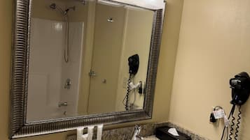 A bathroom with a large mirror, three light fixtures above it, a sink with a granite countertop, a hairdryer, and a fire alarm.