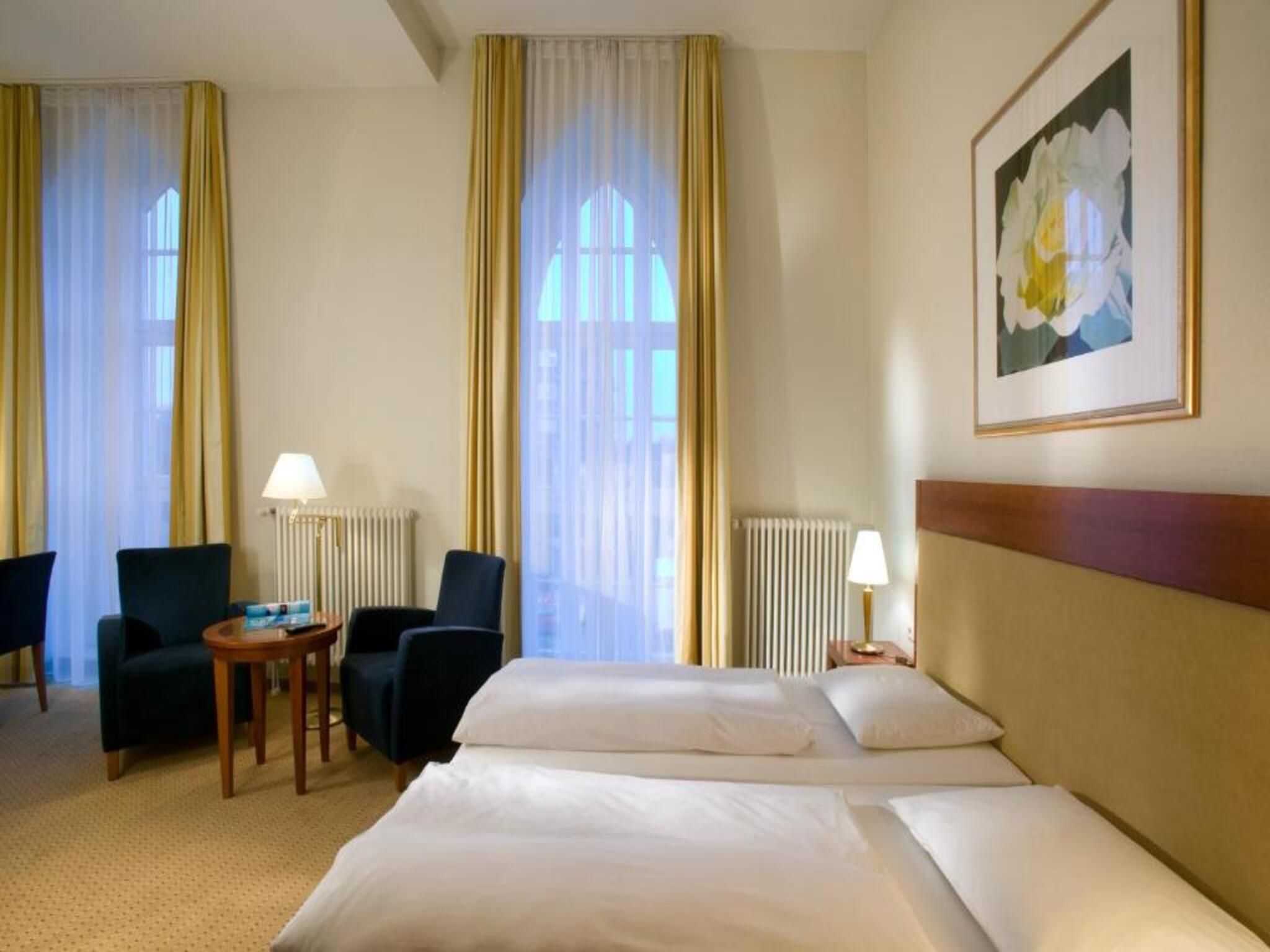 A hotel room with a bed, two chairs, a small table, a radiator, and a window with curtains.