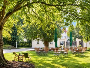 An outdoor seating area with tables and chairs, surrounded by lush greenery and a large tree. -  Sunday Hotel Hildesheim Zentrum (Hildesheim)