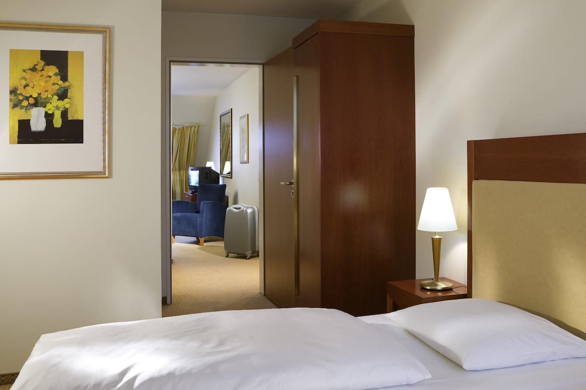 Ein Hotelzimmer mit einem Bett, einem Nachttisch mit Lampe, einem Holzschrank, einem gerahmten Bild an der Wand und Blick in das angrenzende Zimmer.