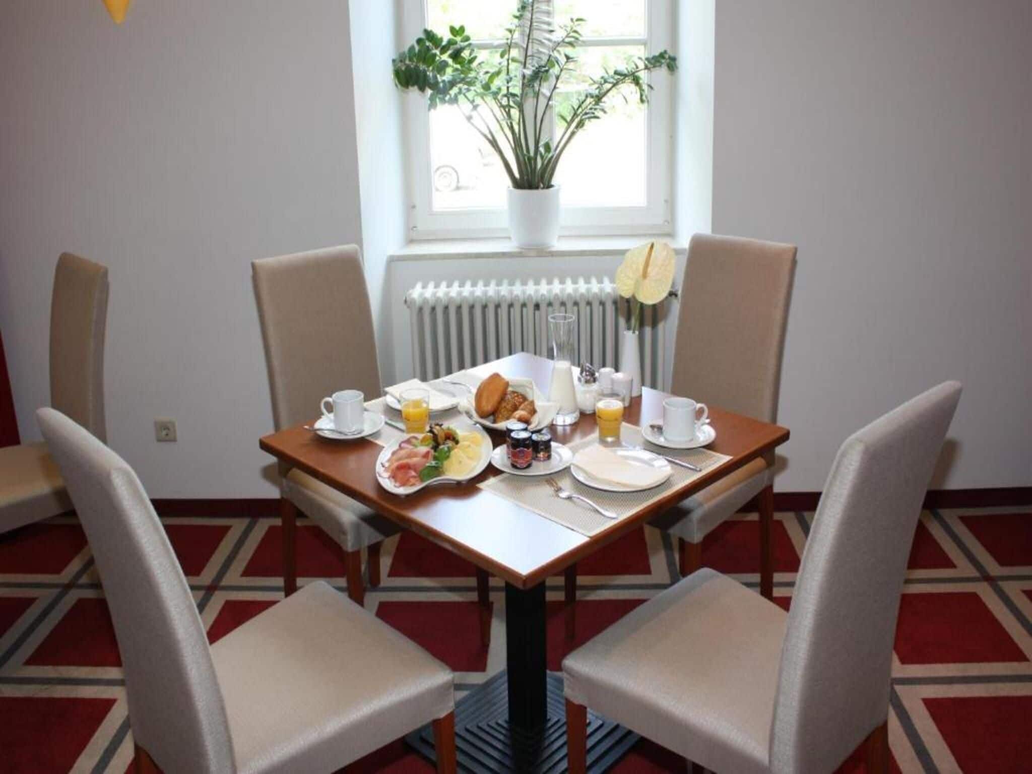 Ein Esstisch, gedeckt für das Frühstück, mit Tellern voller Speisen, Tassen und einem Blumentopf am Fenster.