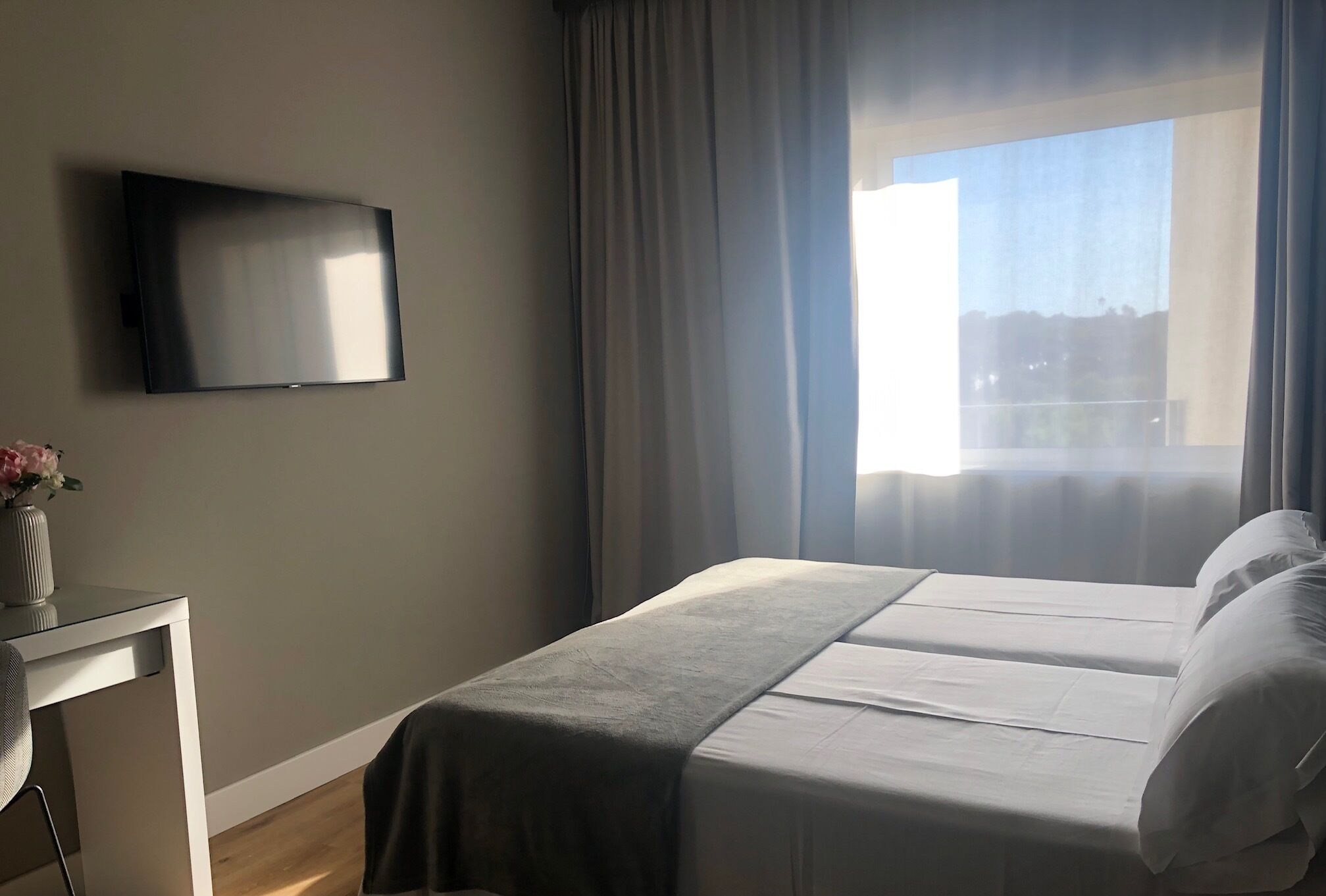 Habitación de hotel con cama, televisor montado en la pared, escritorio con silla y ventana con cortinas.