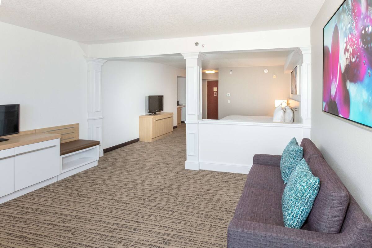 A modern hotel room with a flat-screen TV, a sofa with patterned cushions, a wooden cabinet, and a large abstract painting on the wall.