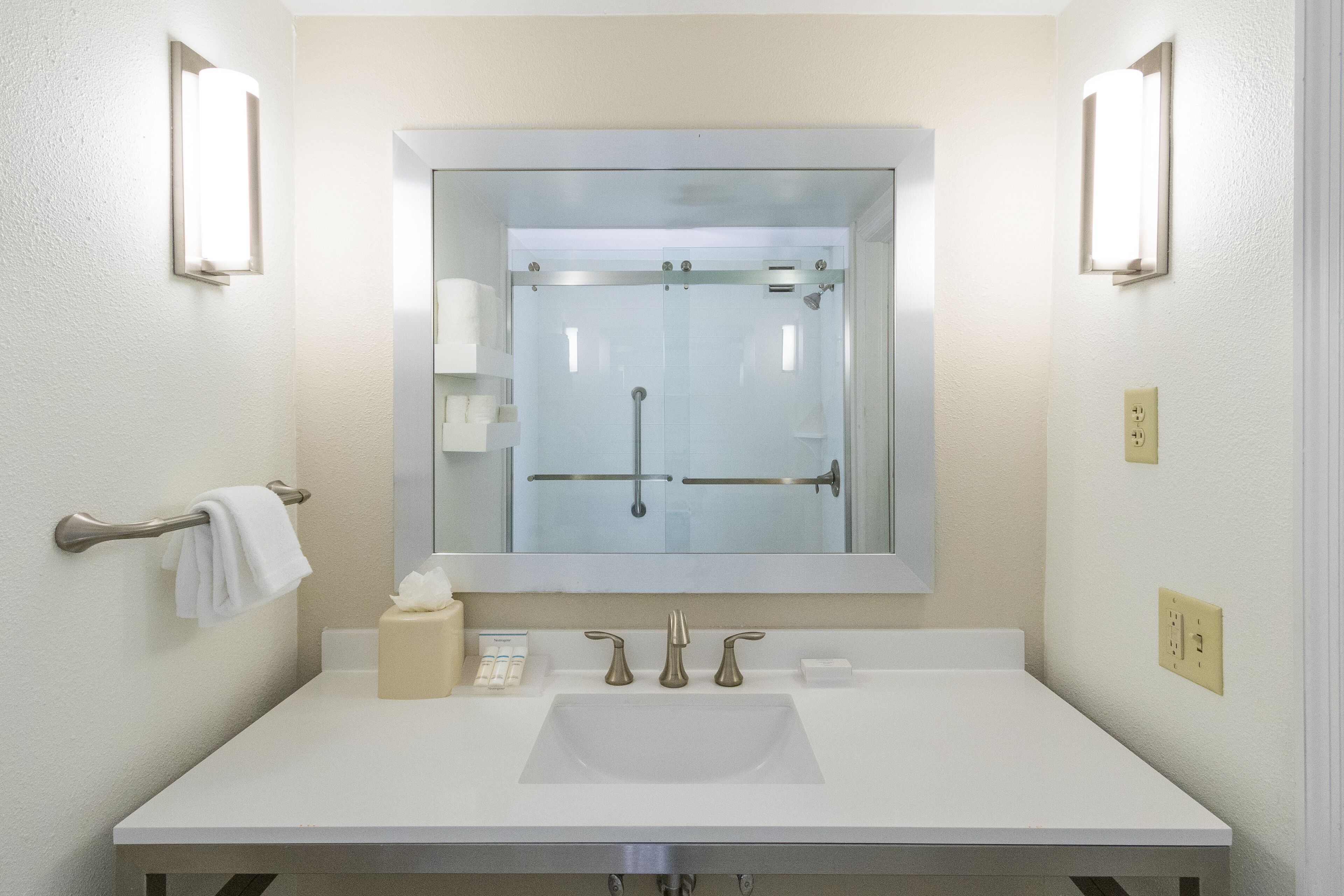 A bathroom with a large mirror, two wall-mounted light fixtures, a towel rack, a soap dispenser, and a glass shower enclosure.