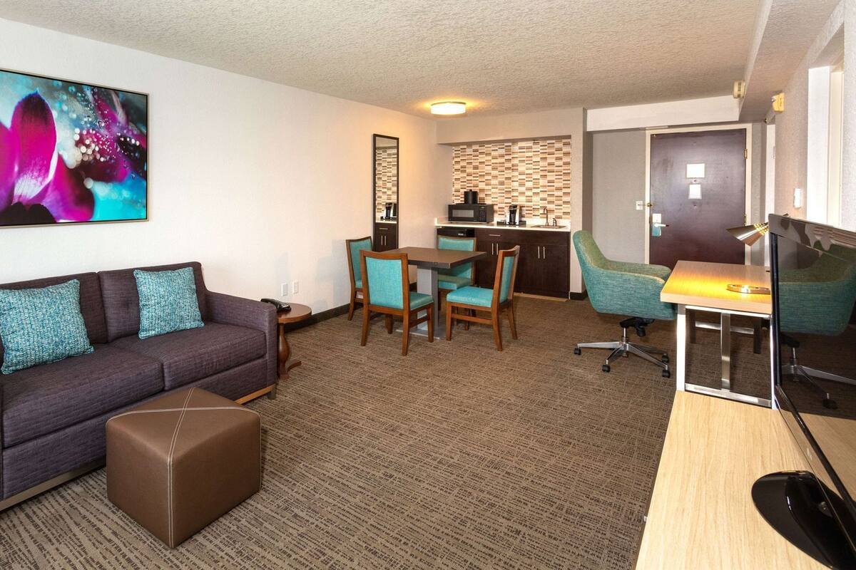 A hotel room with a sofa, ottoman, dining table, chairs, a desk with a computer, a TV, a kitchen area, and a bathroom.