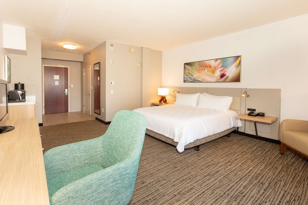 A hotel room with a bed, a desk, a chair, a bedside table, and a painting on the wall.