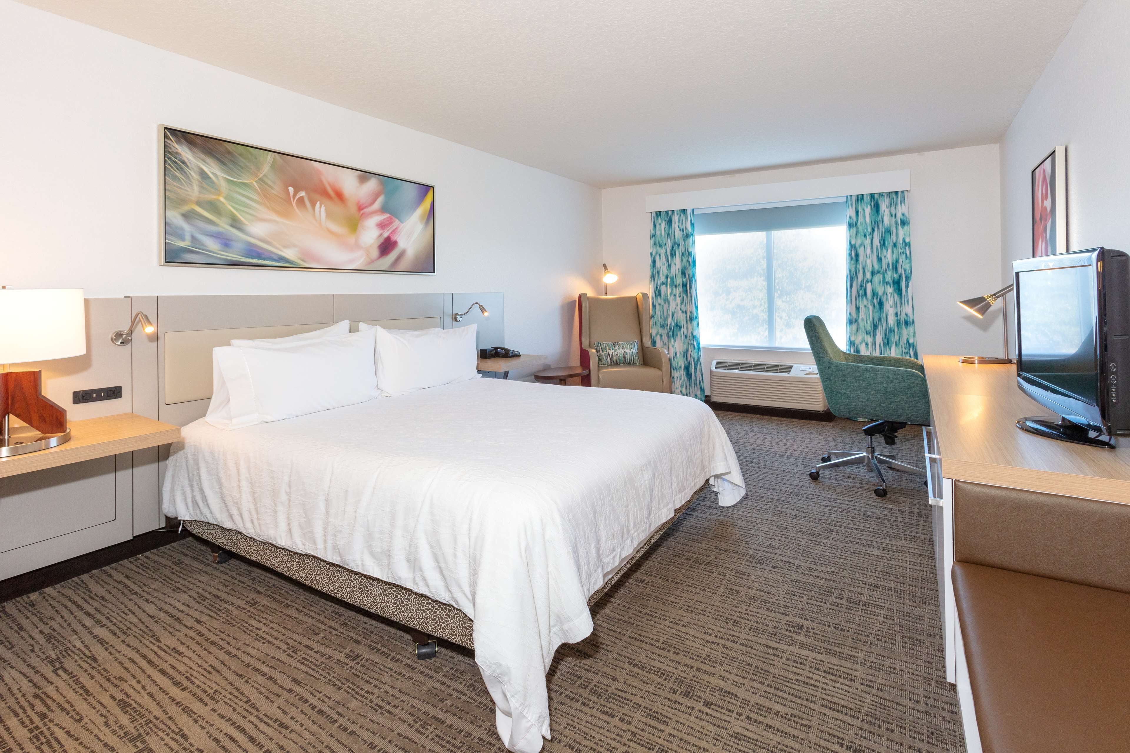 A hotel room with a large bed, a desk with a computer, a chair, a TV, and a window with curtains.