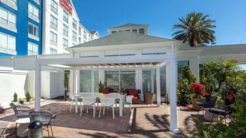 A covered outdoor patio with tables and chairs, a seating area with cushions, and a view of a hotel building in the background.