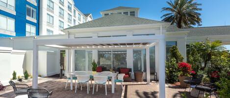 A covered outdoor patio with tables and chairs, a seating area with cushions, and a view of a hotel building in the background.