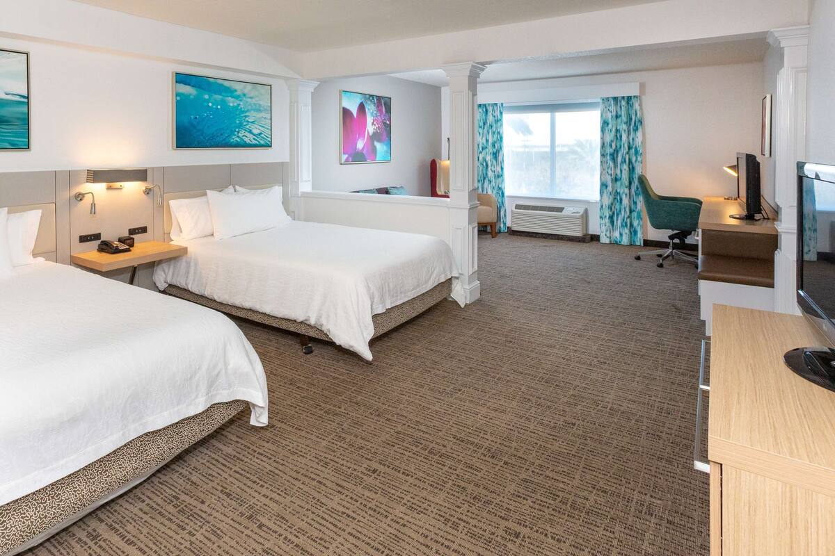 A hotel room with two beds, a desk, a chair, a TV, and artwork on the walls.