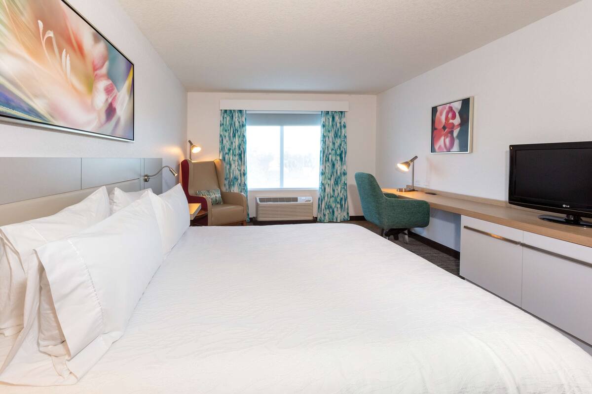 A hotel room with a large bed, a desk with a flat-screen TV, a chair, a lamp, and a window with curtains.