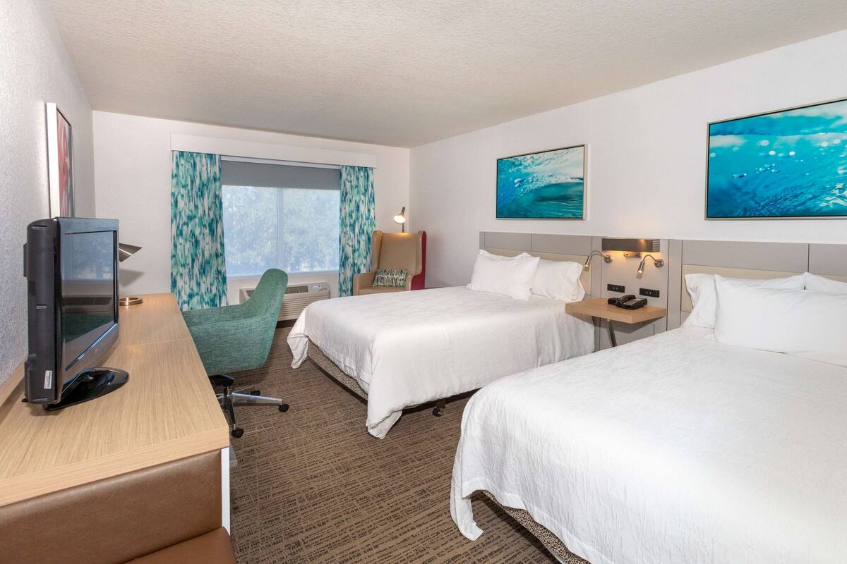 A hotel room with two beds, a desk with a television, a chair, and artwork on the walls.