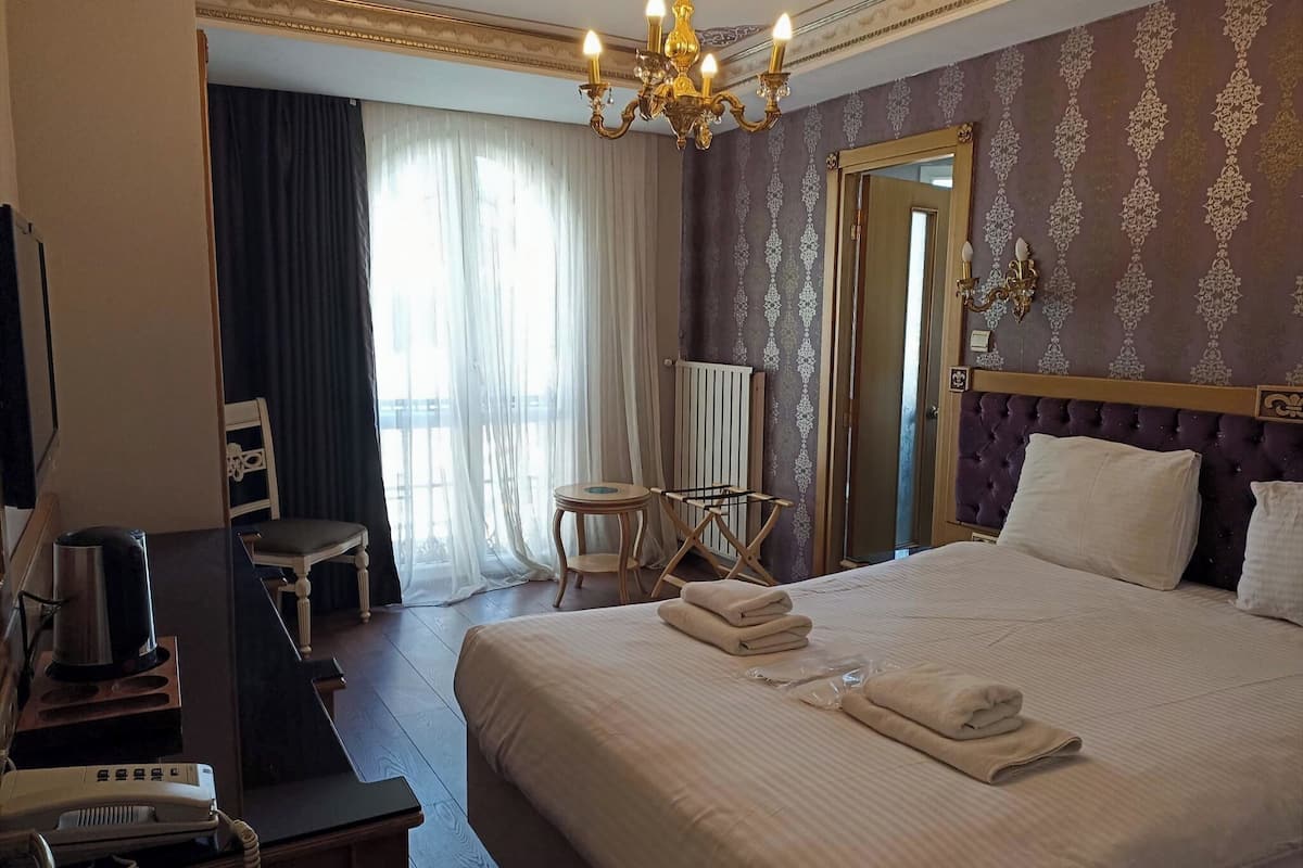A hotel room with a bed, a chair, a desk with a telephone, a radiator, and a chandelier.