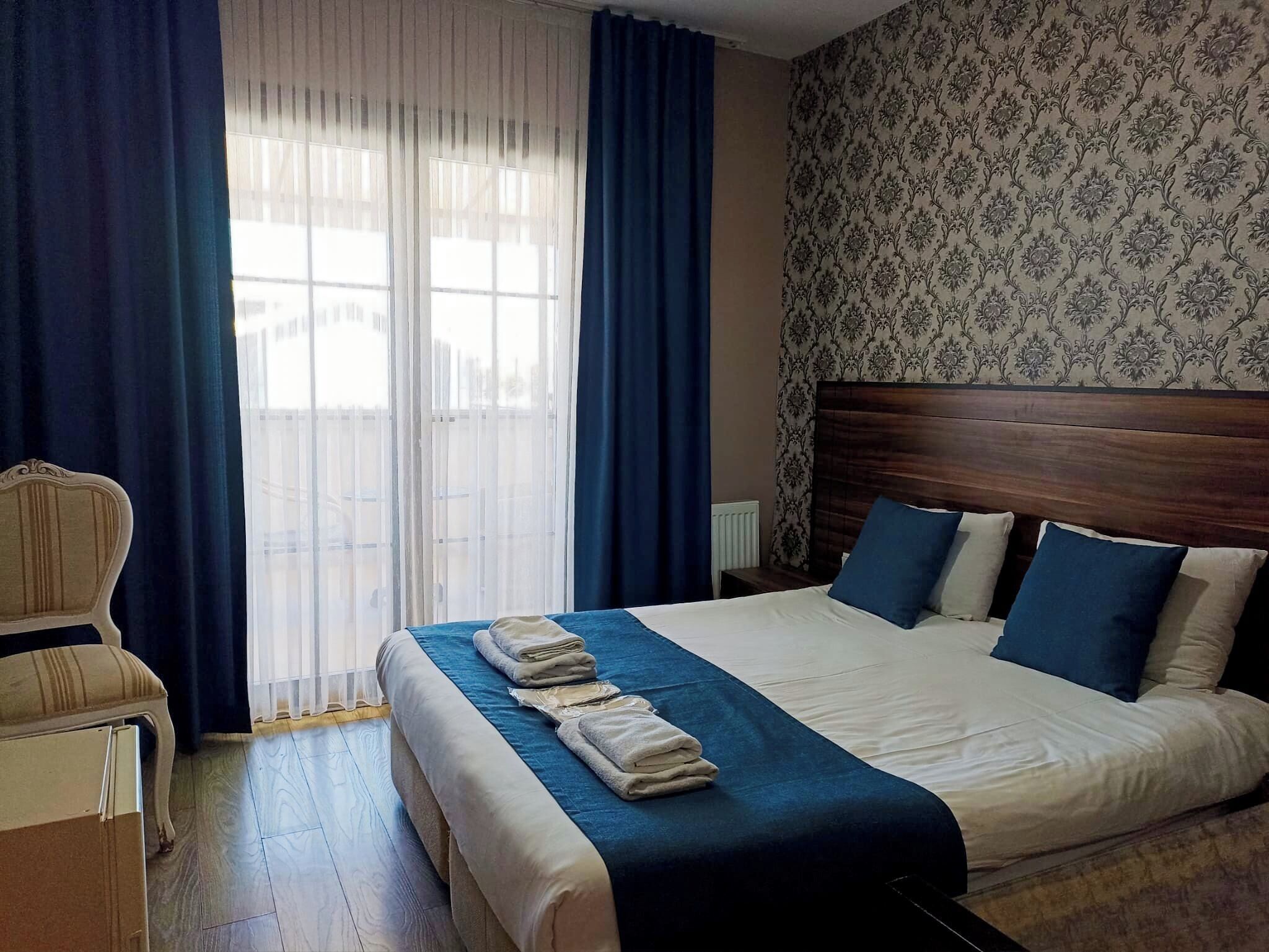 A hotel room with a bed, a chair, a window with curtains, and a patterned wallpaper.