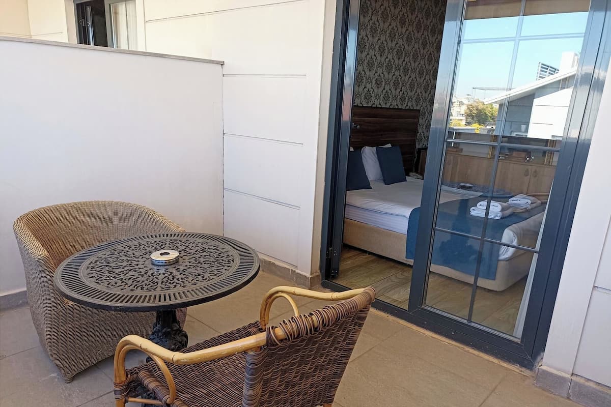 A balcony with a wicker table and chairs, a bed with a blue bedspread, and a view of buildings through a glass door.