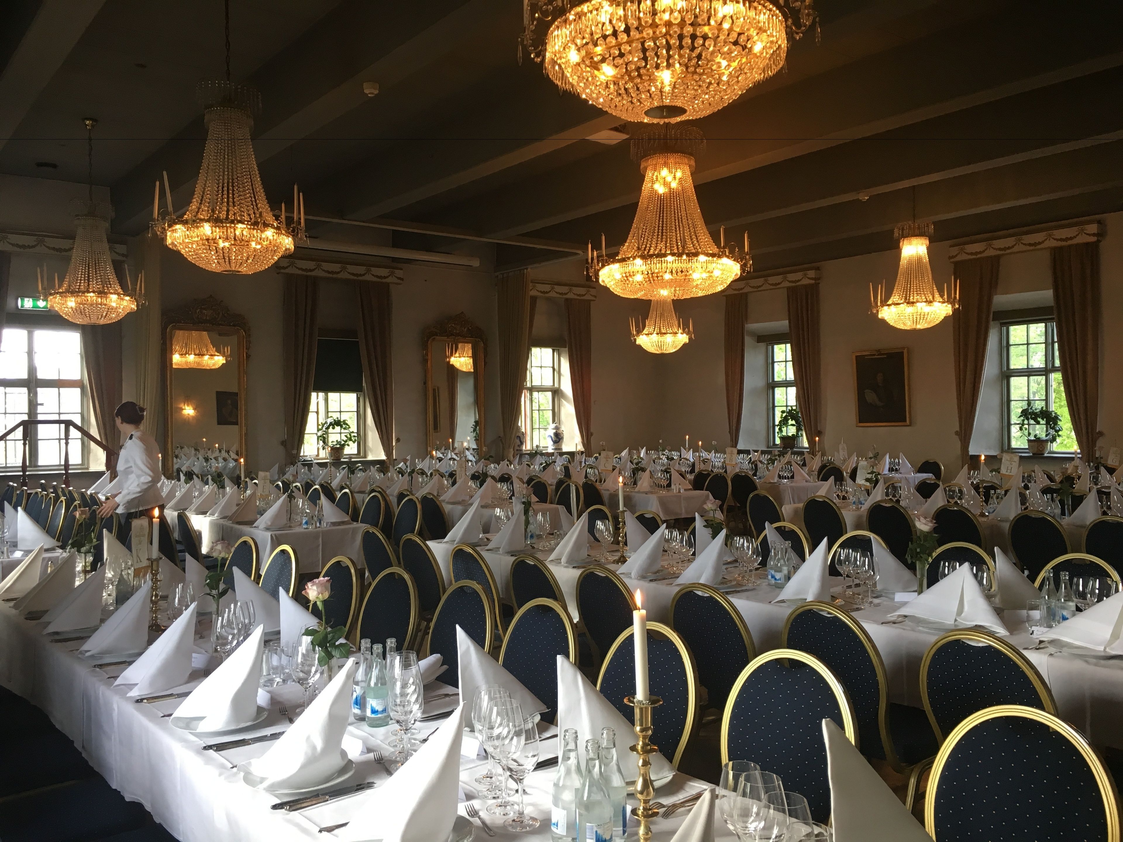 Een balzaal met meerdere gedekte tafels voor een formeel evenement, met kroonluchters en een grote spiegel.