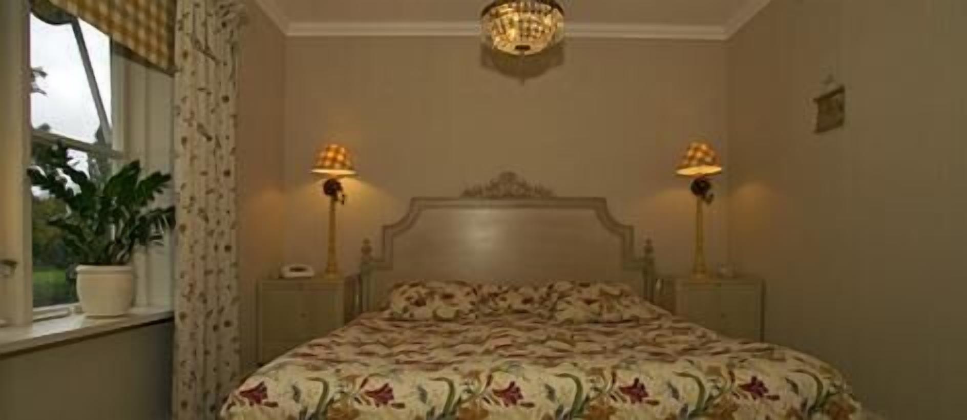 A bedroom with a large bed, two bedside lamps, a chandelier, and a window with curtains.