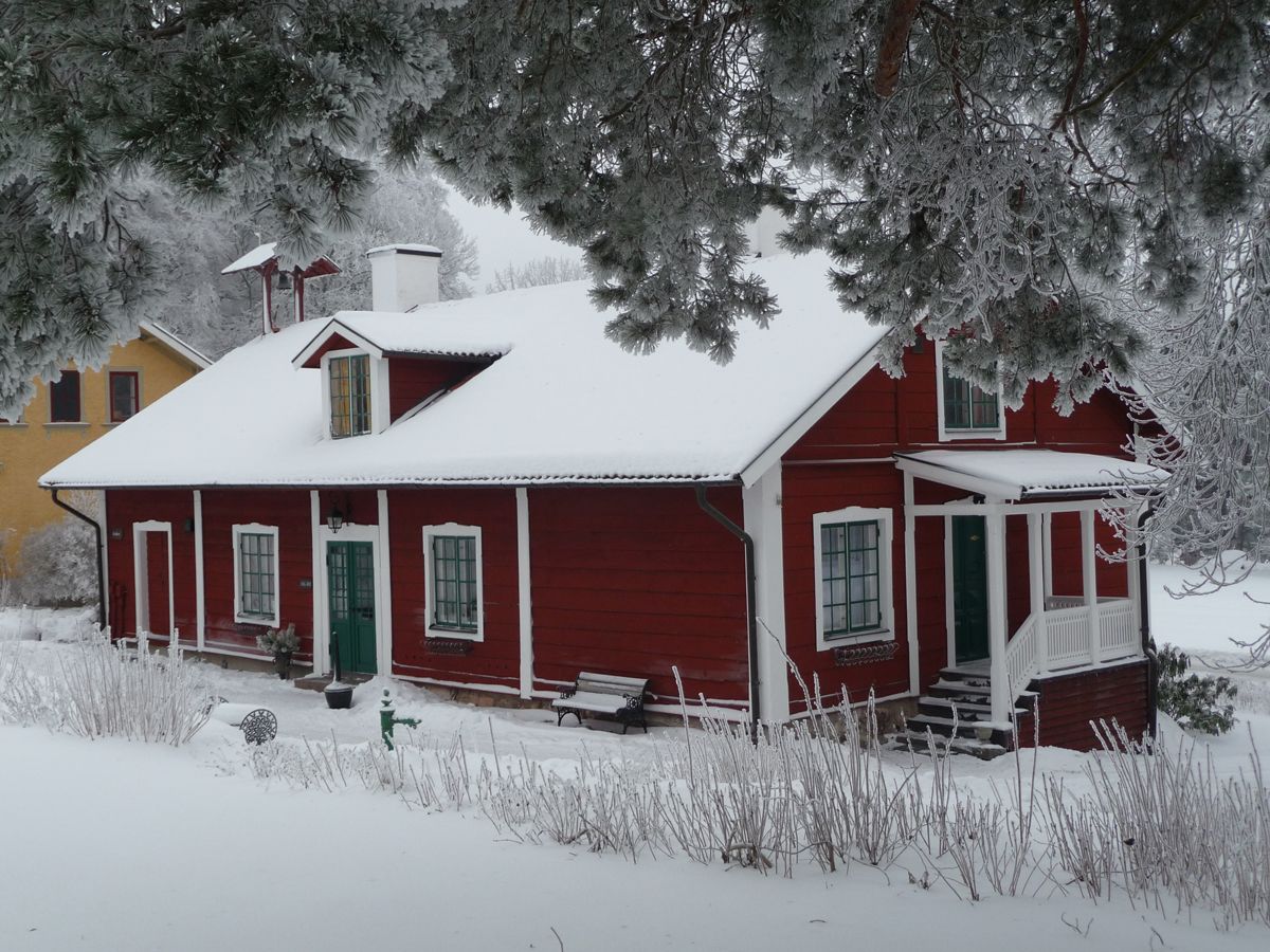 Una casa de madera roja con un porche, rodeada de nieve en el suelo y árboles.