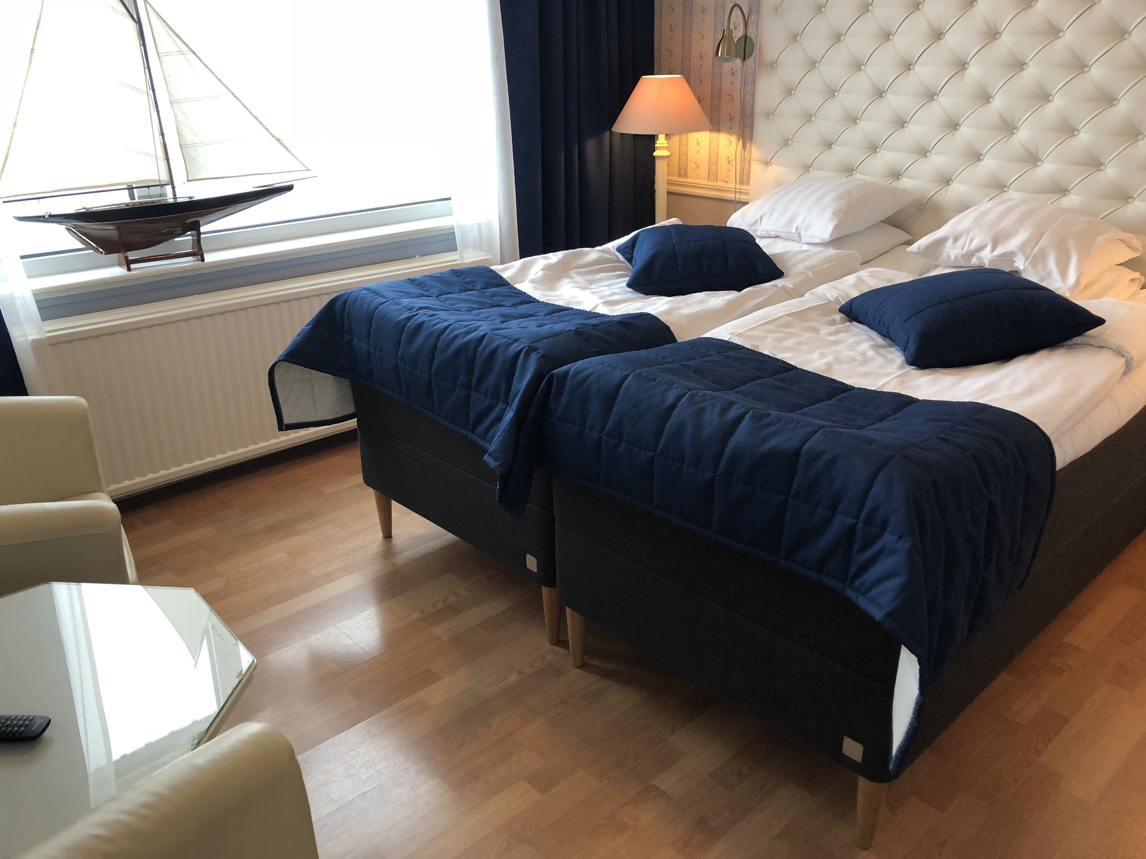 A hotel room with a large bed, two pillows, a bedside lamp, a window with a sailboat model, a chair, and a small table.
