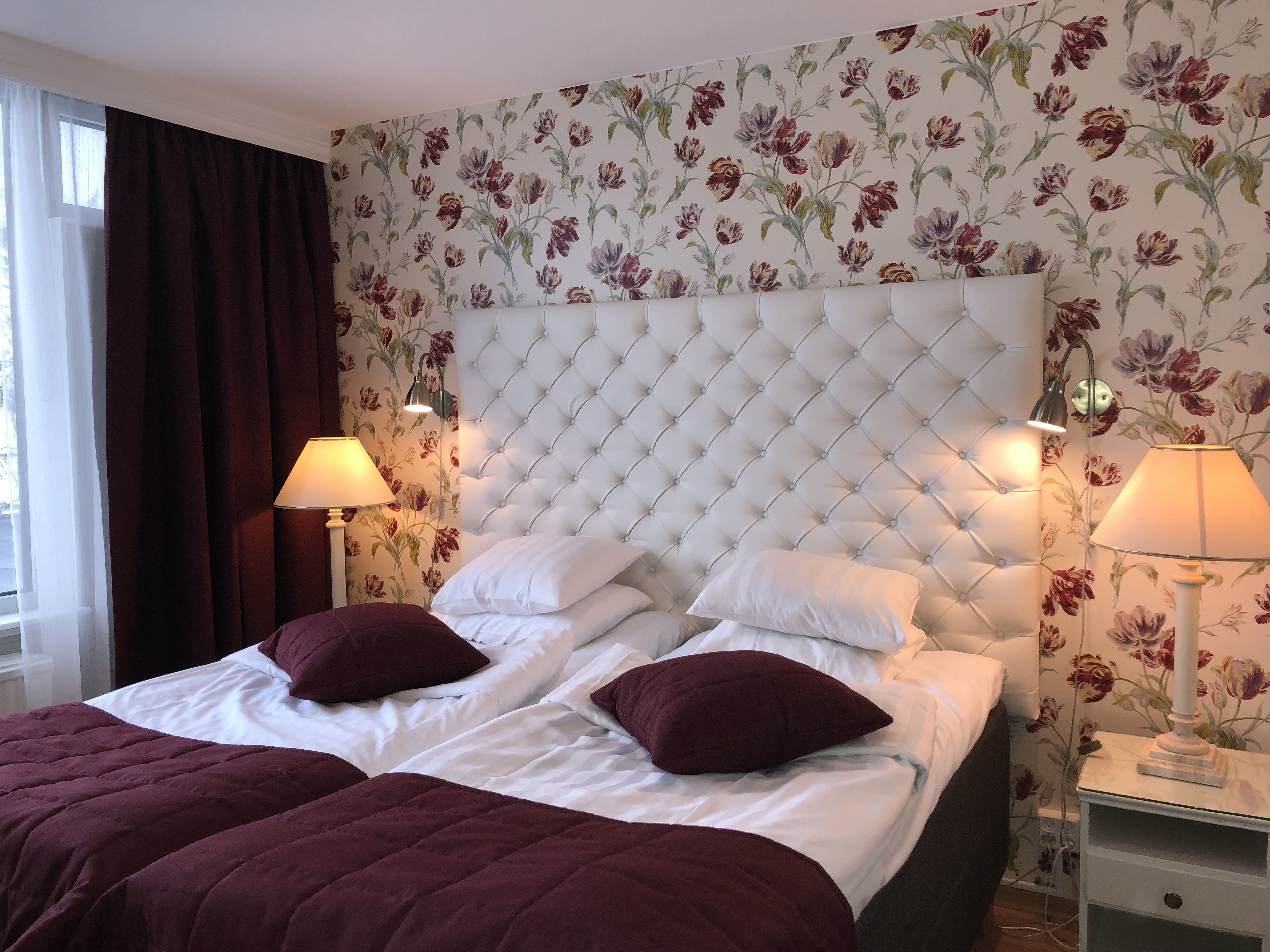 Une chambre d’hôtel avec une tête de lit capitonnée, deux lits avec une literie blanche et bordeaux, un papier peint à motifs floraux et deux lampes de chevet.