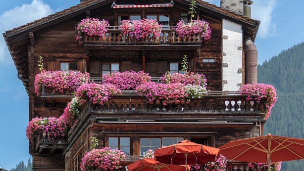 A traditional wooden building with balconies adorned with pink flowers, a sign reading "Chesa Arischuna," and a mountain backdrop. - Hotel Chesa Grischuna (Klosters-Serneus)