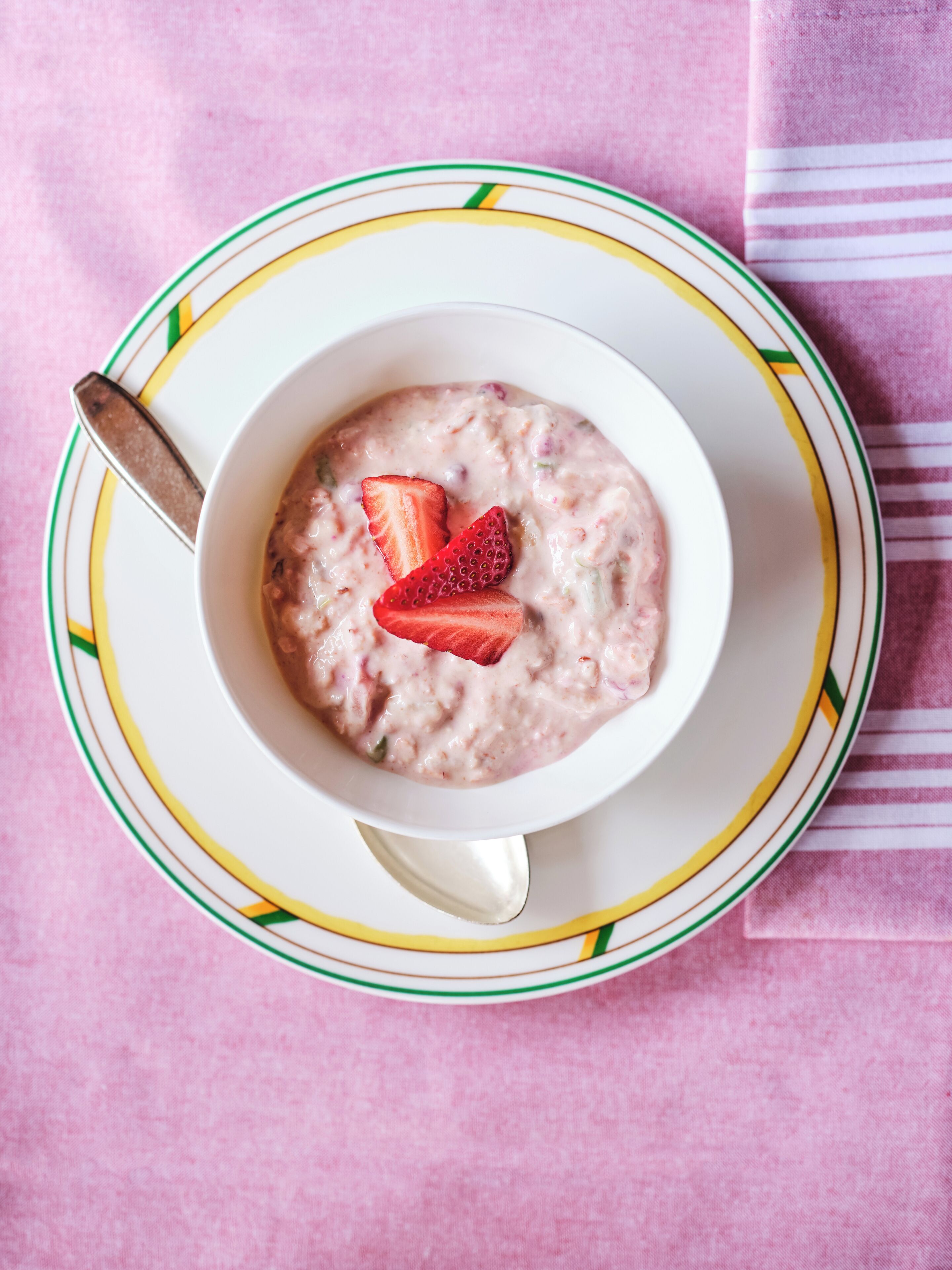 Una ciotola di porridge con fragole su un piatto bianco con bordo verde e giallo, posata su un tessuto rosa.