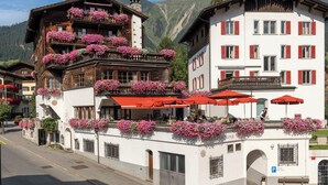 A traditional European building with balconies adorned with flowers, a street in front, and a mountain backdrop. - Hotel Chesa Grischuna (Klosters-Serneus)