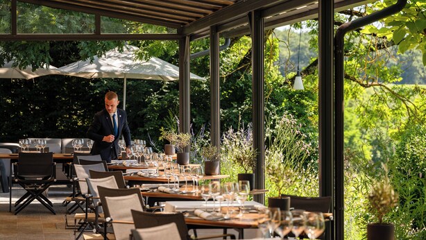 Un área de comedor al aire libre con mesas dispuestas para una comida, rodeada de ventanas amplias y plantas en macetas.
