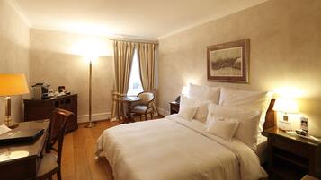 Habitación de hotel con cama, escritorio, silla, lámpara, mesita de noche y un cuadro en la pared.