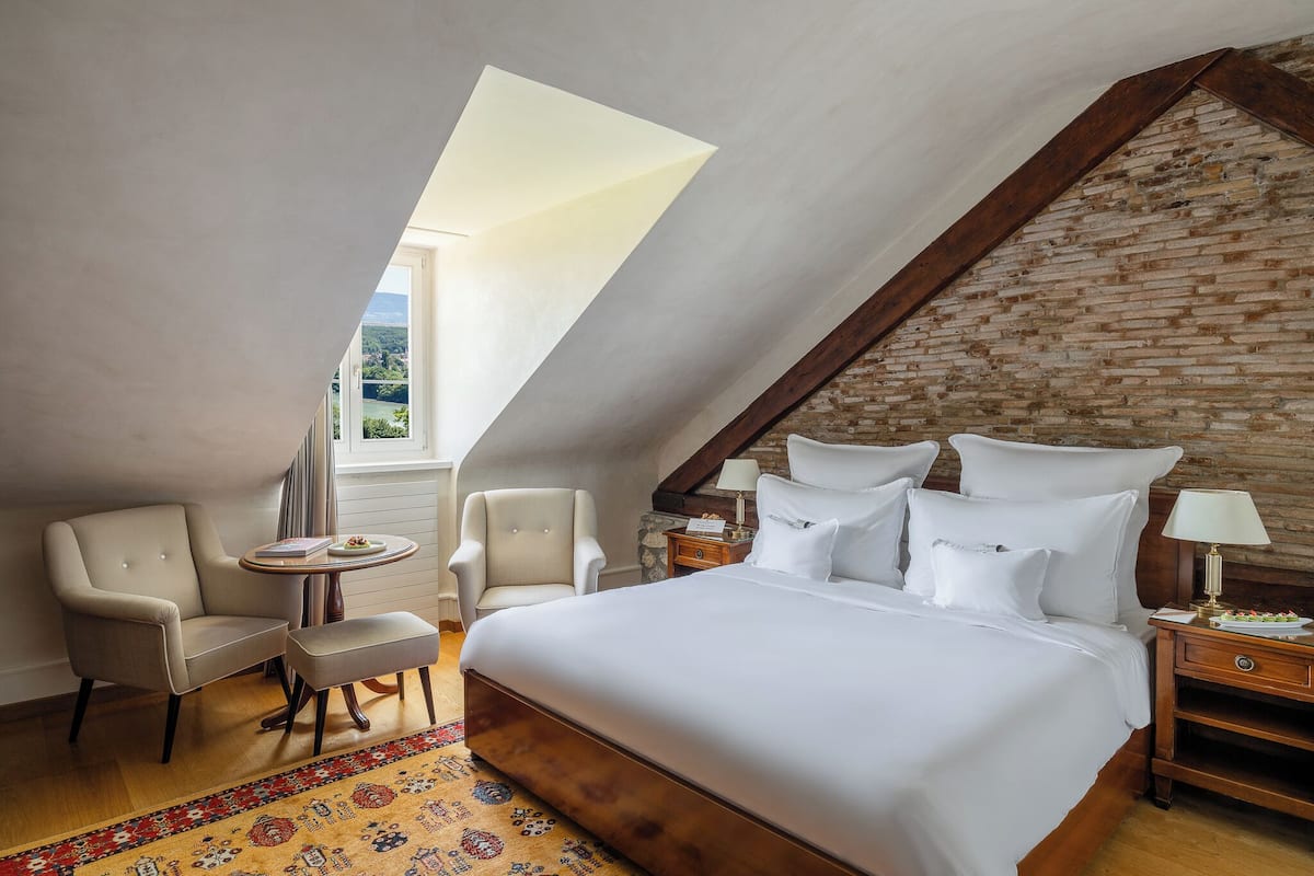 Une chambre avec un grand lit, deux fauteuils, une petite table ronde et une table de chevet avec une lampe. La pièce a un plafond en pente et un mur en briques.