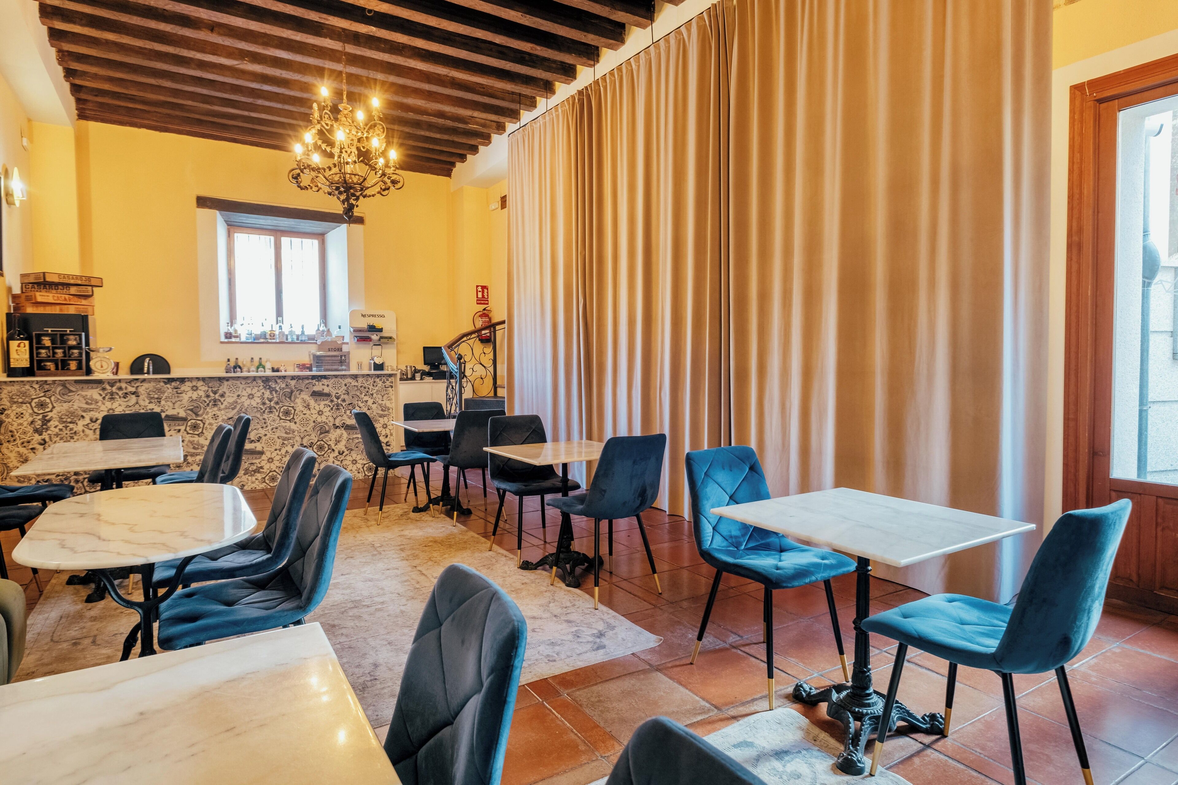 A restaurant with marble-topped tables and blue upholstered chairs, a chandelier, and a window with curtains.