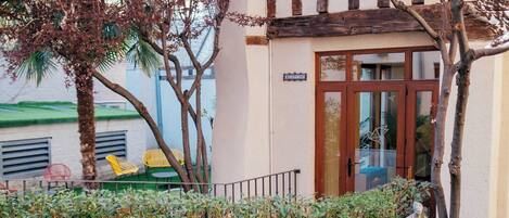 Un espacio al aire libre con sillas amarillas y una mesa, rodeado de follaje y un edificio con puerta de madera.