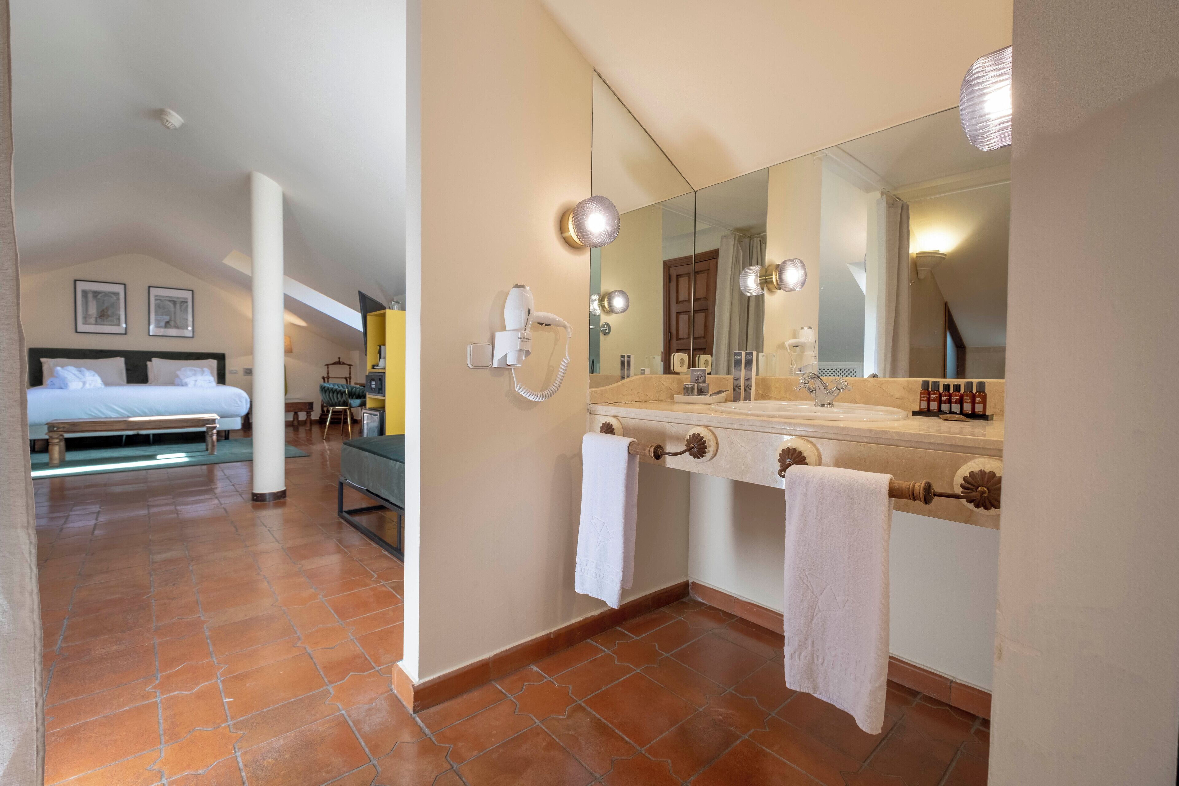 A hotel room with a bed, a bathroom with a large mirror, a hairdryer, and a towel rack.