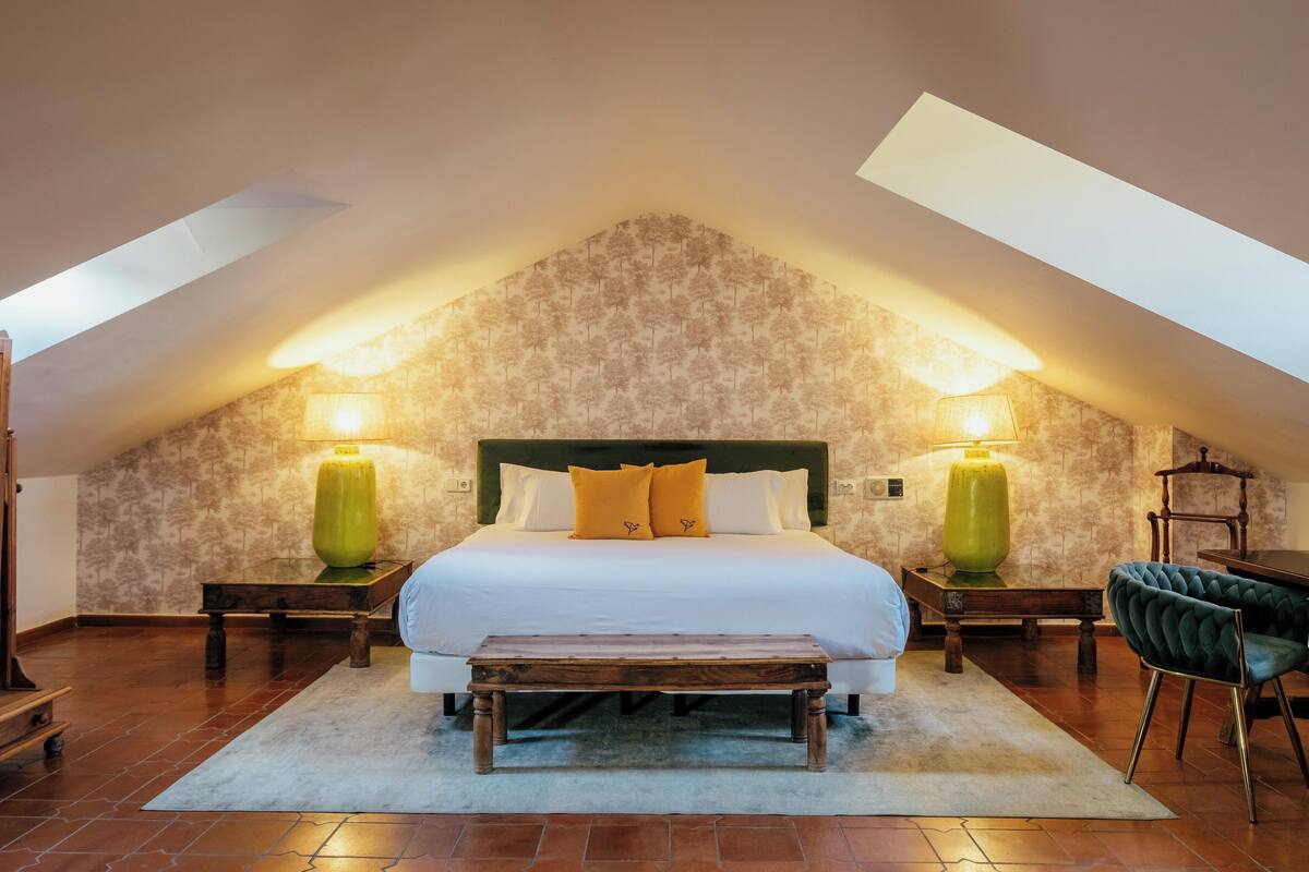 Ein Schlafzimmer mit schrägverdeckter Decke, einem großen Bett mit weißer Bettwäsche und zwei gelben Kissen, zwei Nachttischlampen, einer hölzernen Bank und einem grünen Polsterstuhl.