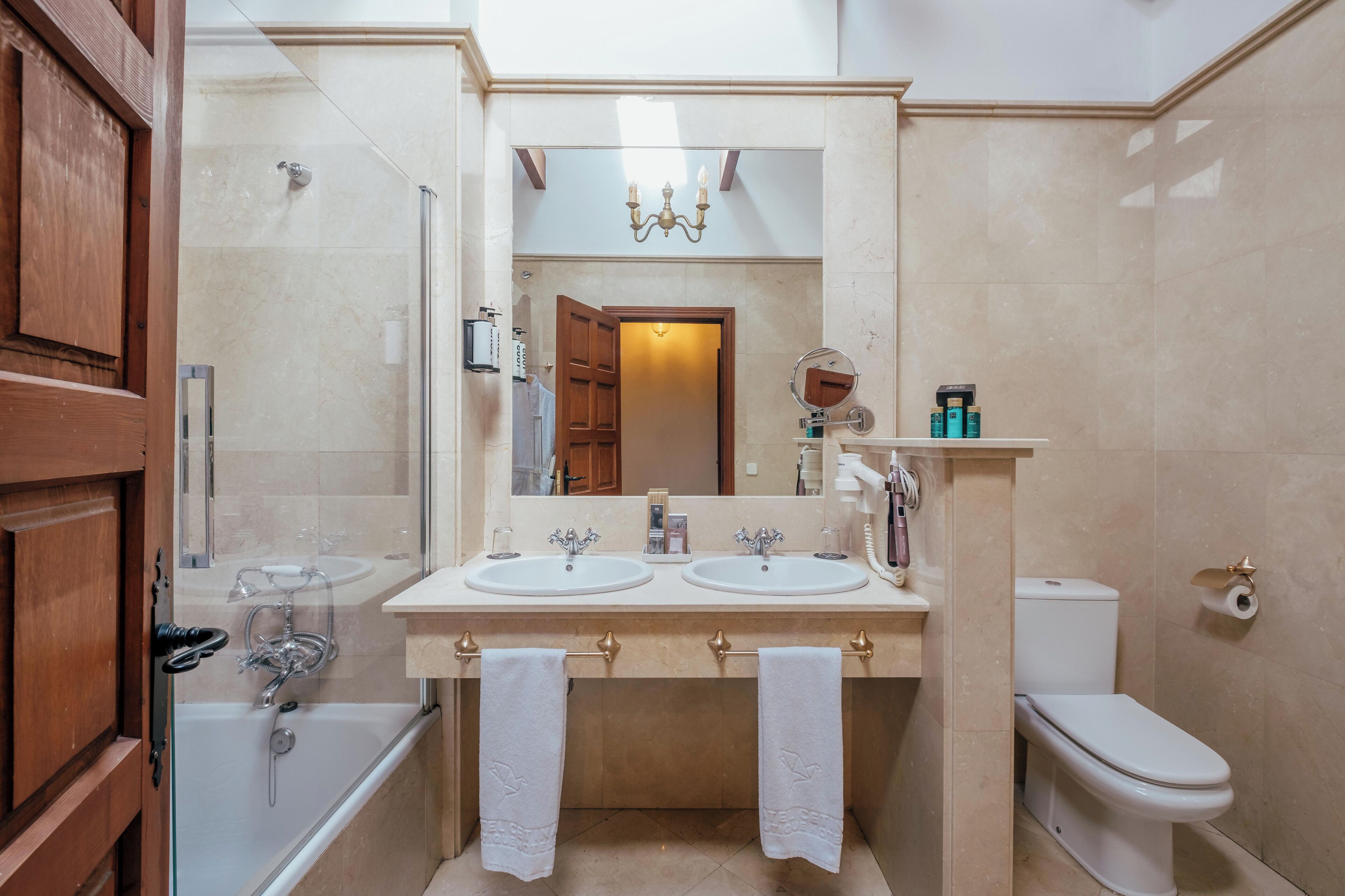 Baño con dos lavamanos, inodoro, cabina de ducha de vidrio y espejo con candelabro.