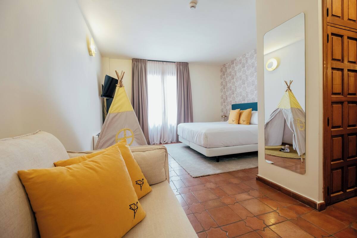 Ein Hotelzimmer mit einem Bett, einem Sofa mit gelben Kissen, einem Spiegel, einem Fenster mit Vorhängen und einer Holztür.