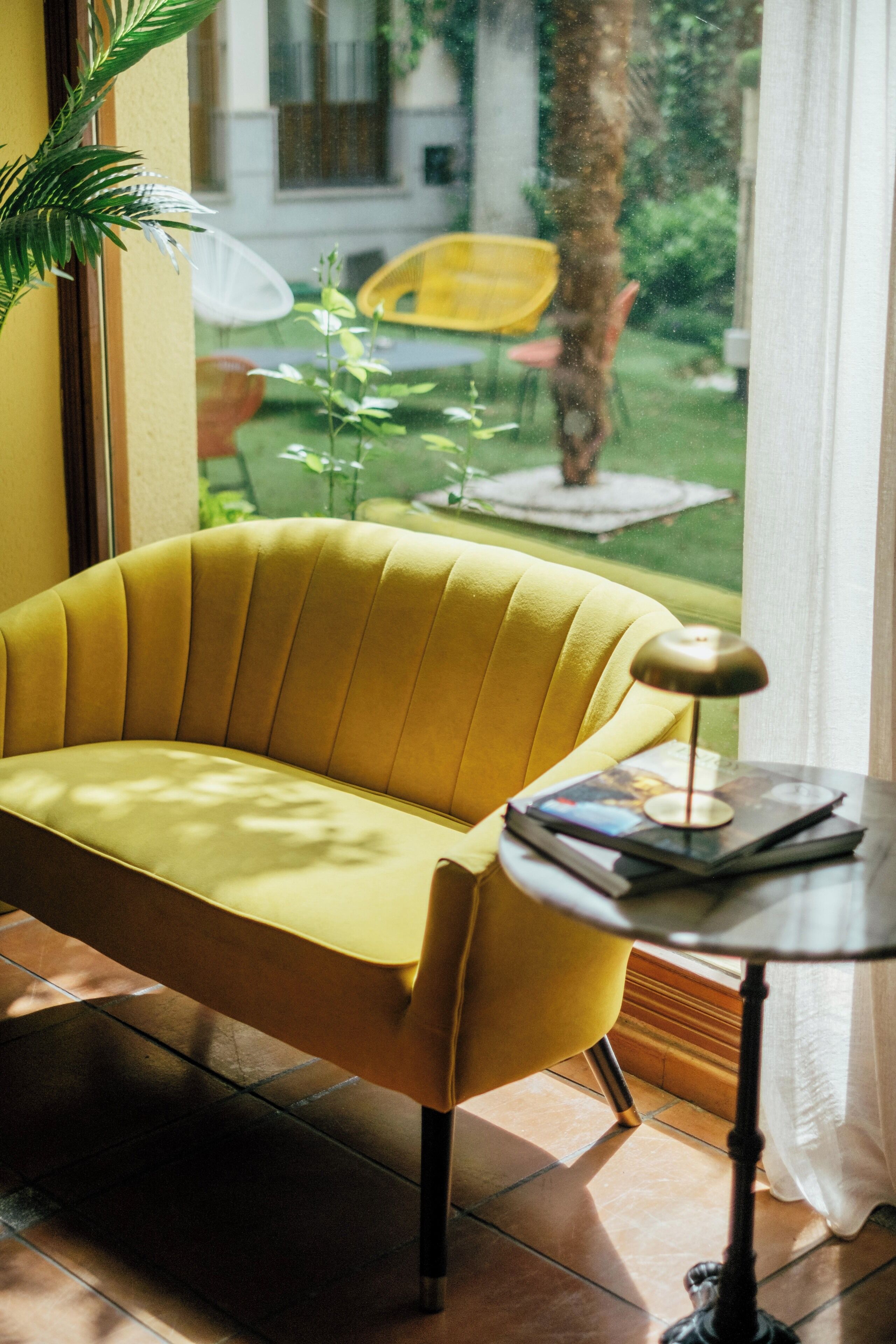 Un sofá amarillo, una mesita con una lámpara, y vistas a un jardín con una silla amarilla y una planta.