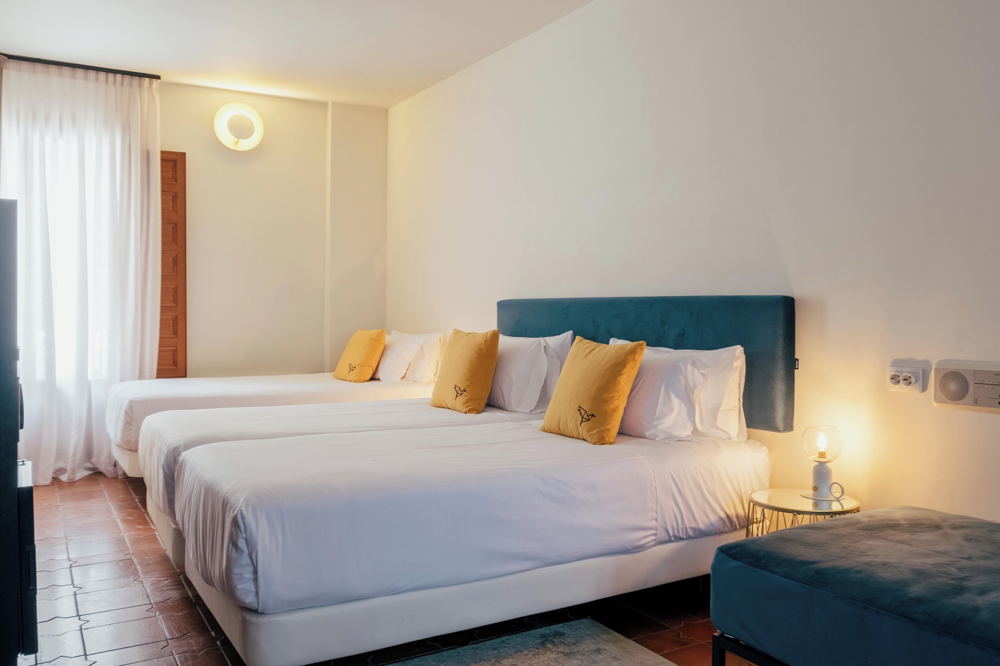 Une chambre d’hôtel avec deux lits, un applique murale ronde, une petite table avec une lampe et un pouf bleu.