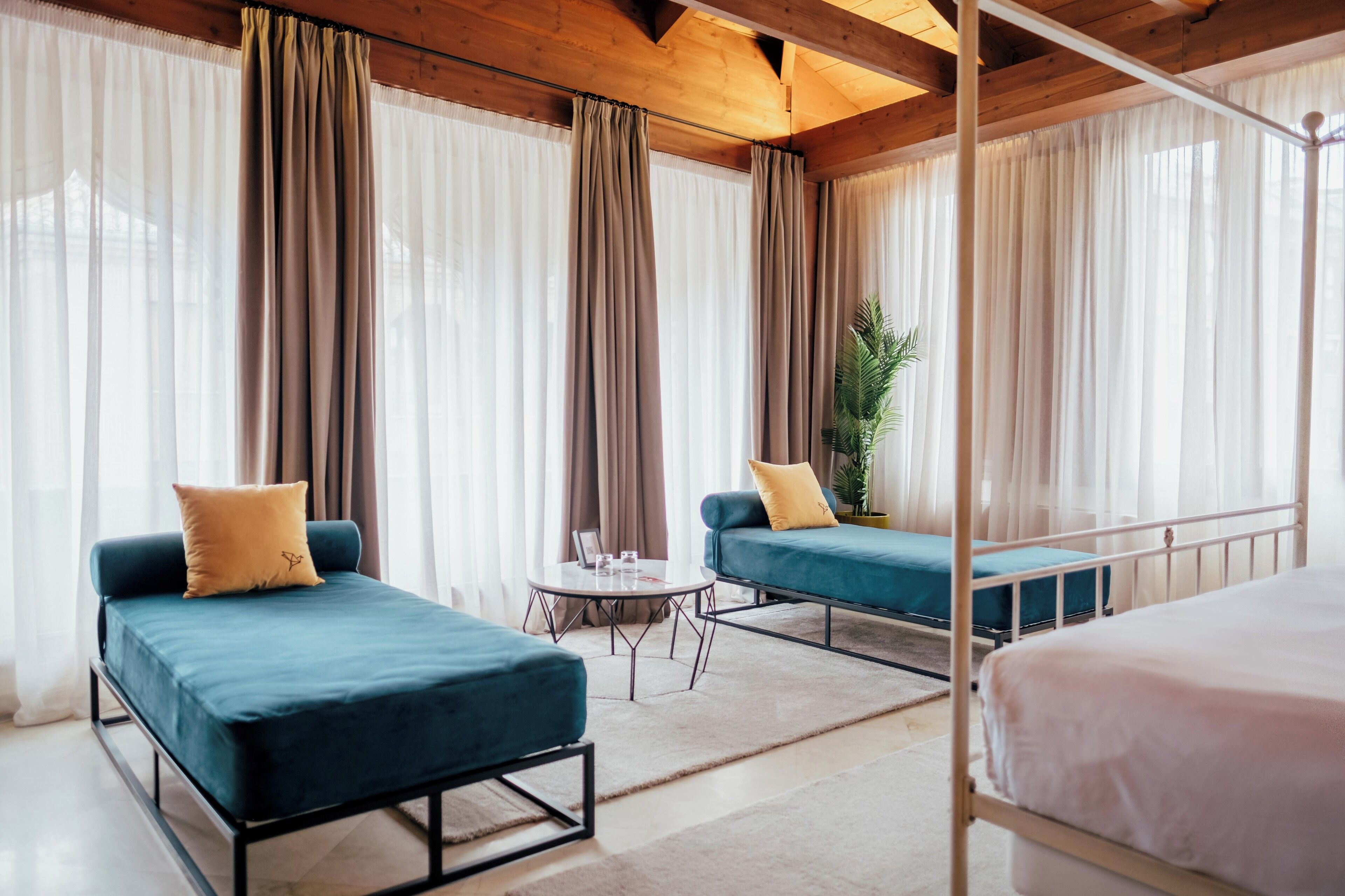 A modern hotel room with two single beds, a small round table, and two blue sofas with cushions.