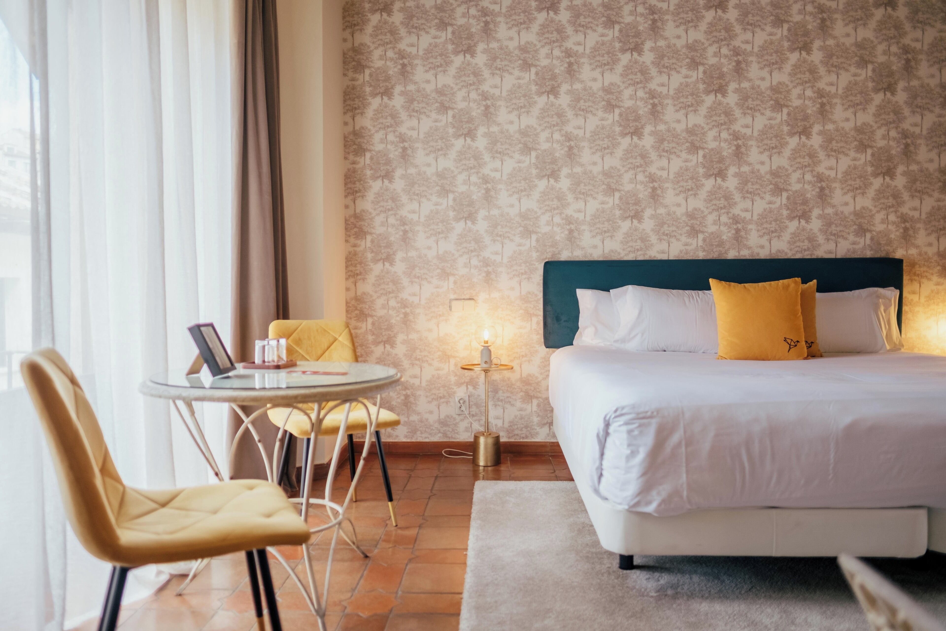 A hotel room with a bed, a round table with chairs, a lamp, and a window with curtains.
