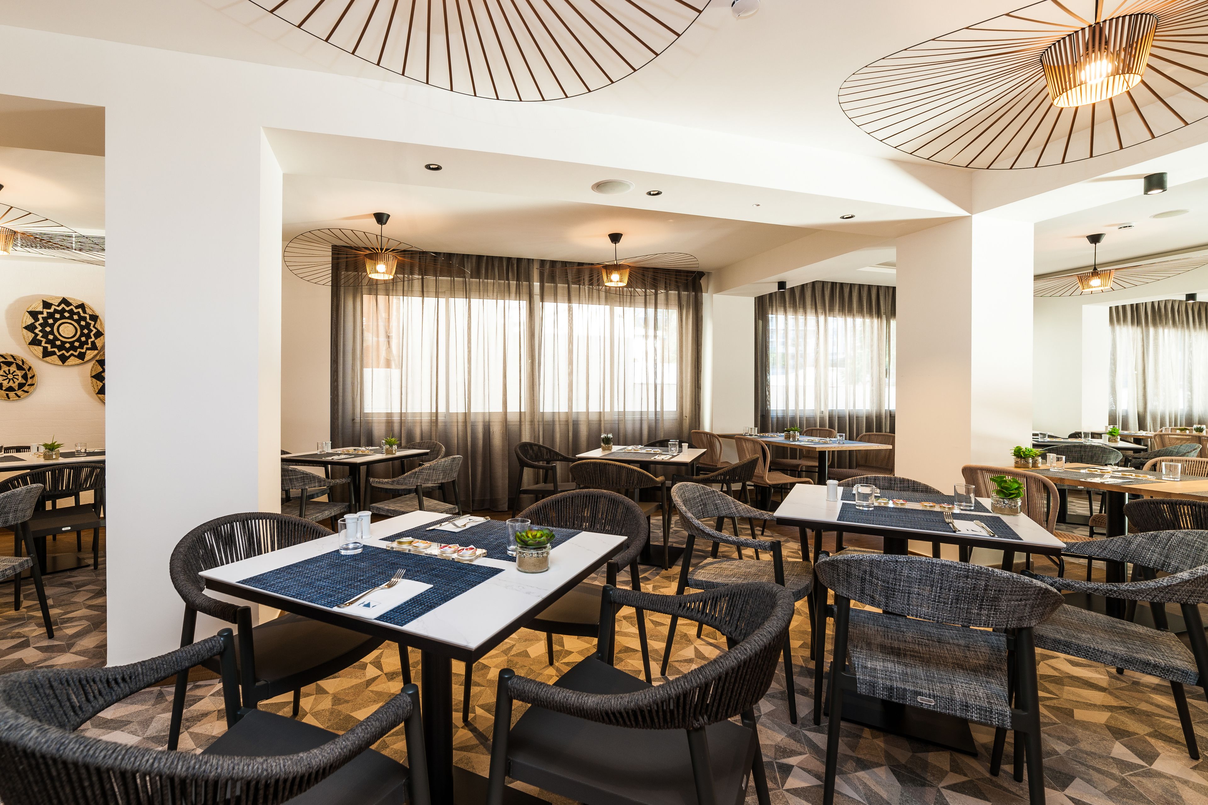 Un comedor de hotel con varias mesas y sillas, amplias ventanas con cortinas e iluminación de techo.