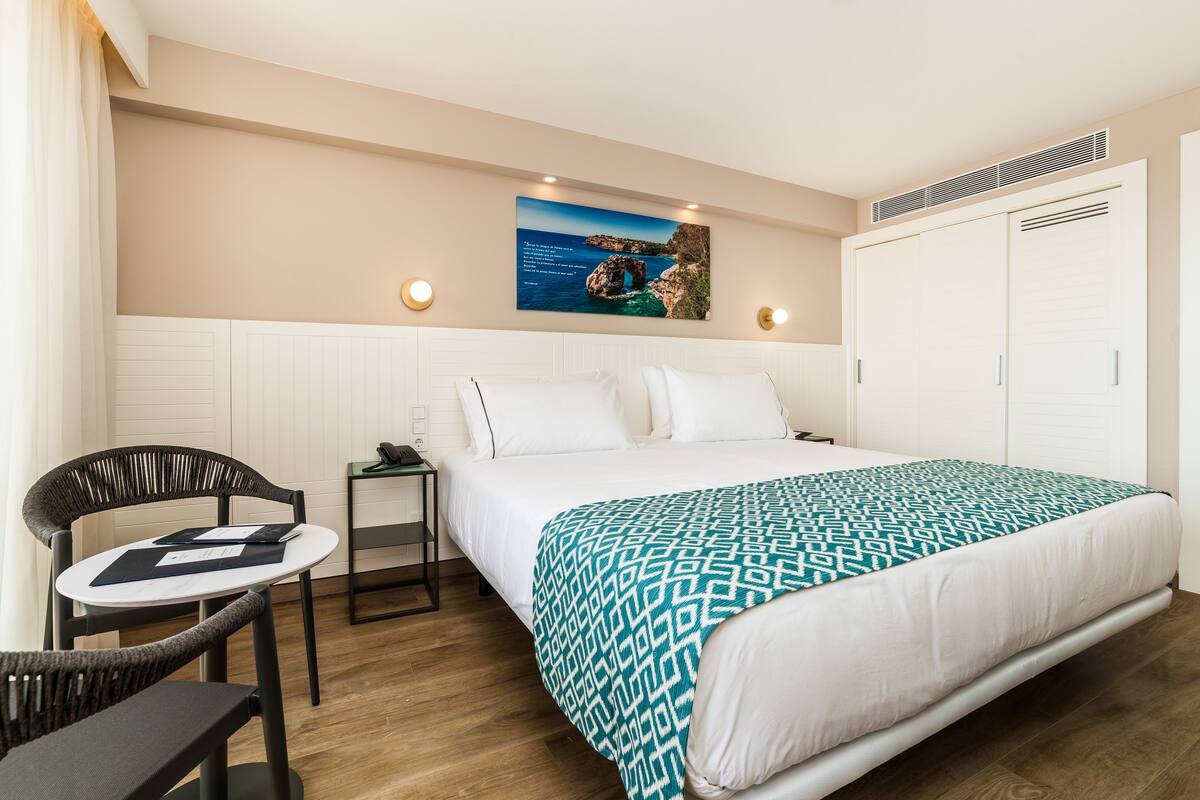 A hotel room with a bed, a chair, a small table, a nightstand, and a painting on the wall.