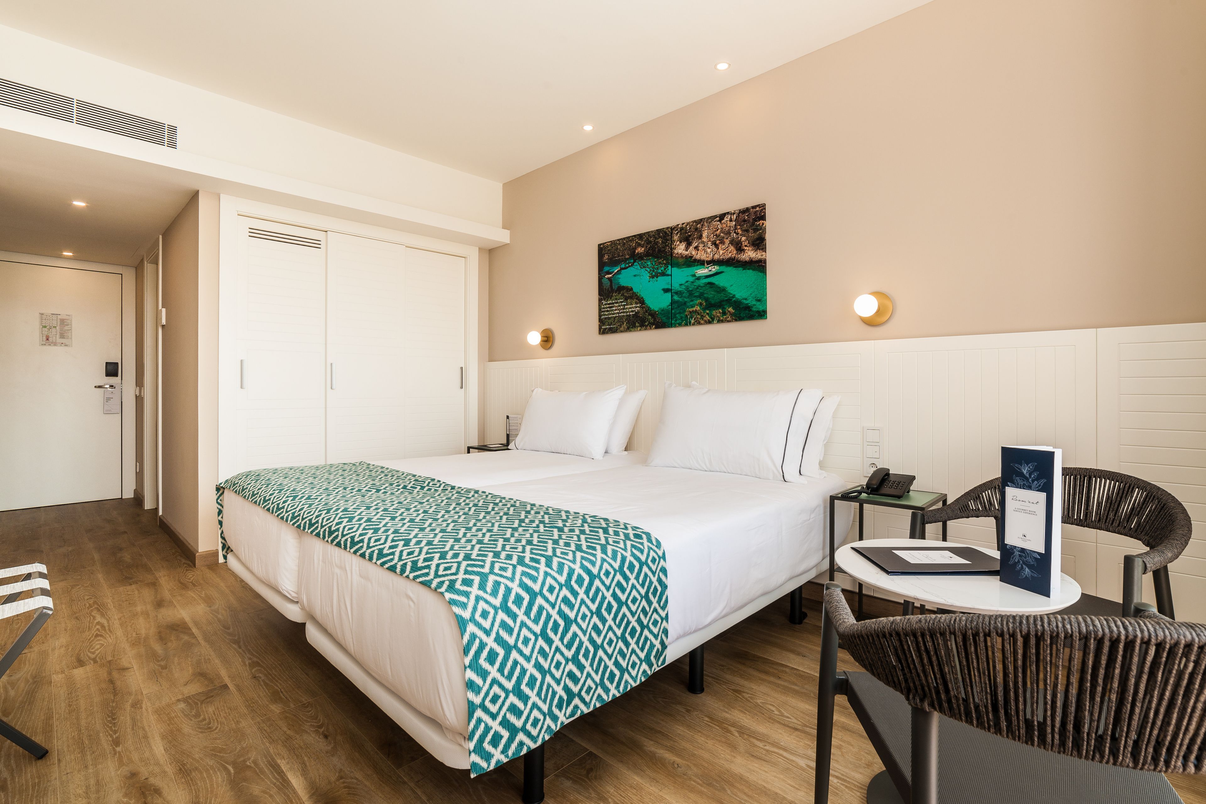 A hotel room with a bed, a chair, a small table, and a painting on the wall.