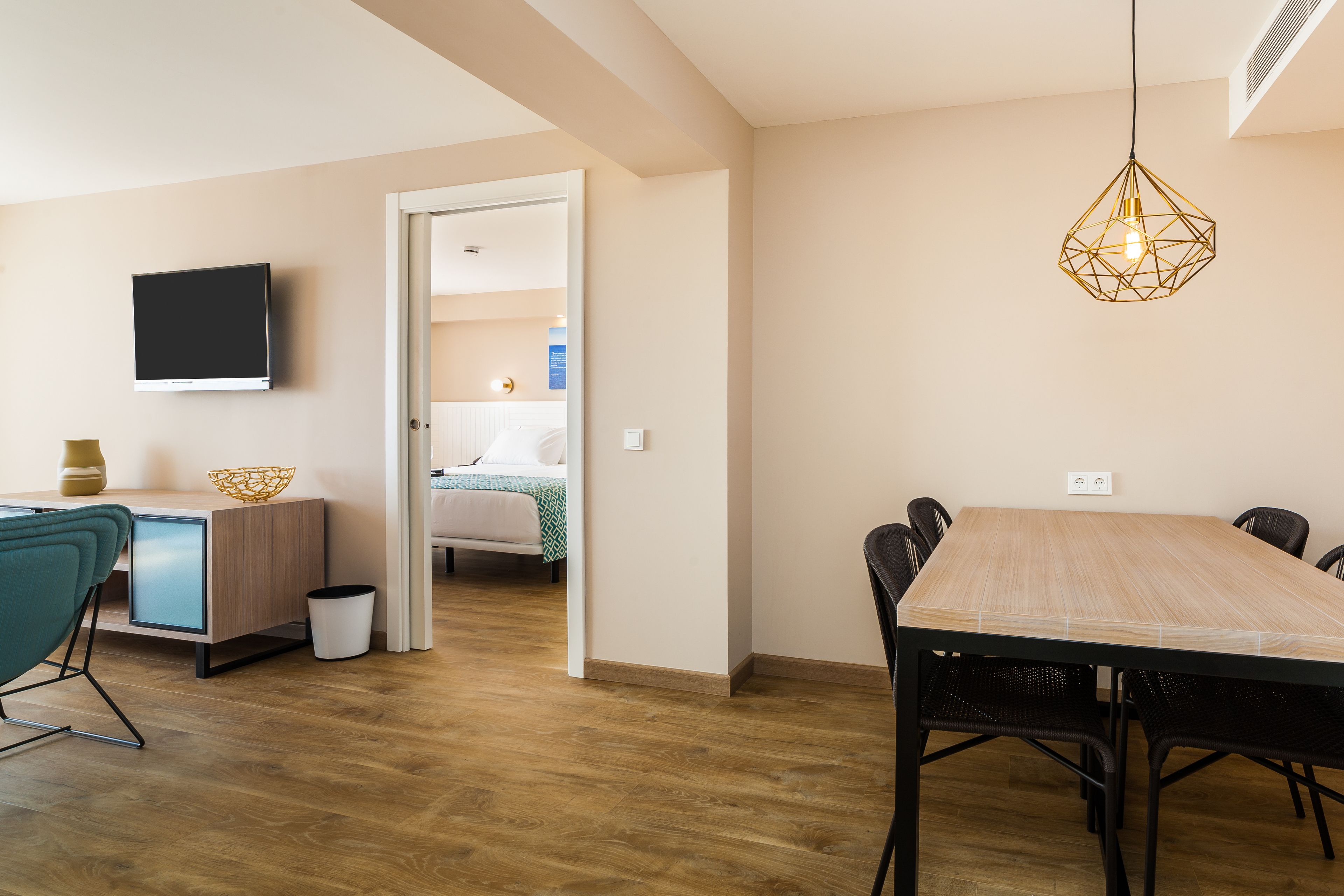Une chambre d’hôtel moderne avec une table à manger en bois, un téléviseur fixé au mur et un luminaire suspendu au design géométrique.